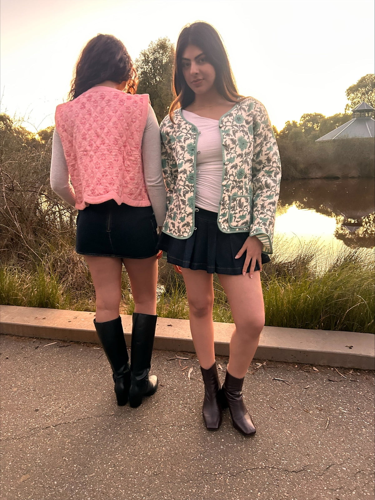 Two women standing outdoors near a pond at sunset, wearing patterned and solid outerwear. One woman is shown from the back wearing a pink patterned vest over a light gray top and black skirt, while the other woman stands facing forward wearing a sage green and white floral patterned bomber jacket over a white top and black pleated skirt.